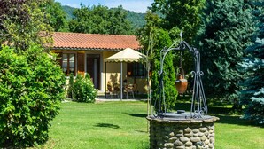 Un puits en pierre avec une gouttière en métal, entouré d’une végétation luxuriante, et une maison avec un toit en tuiles en arrière-plan.