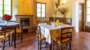 Un coin repas avec une cheminée, des chaises en bois, une table mise pour le petit-déjeuner et un grand tableau religieux accroché au mur.