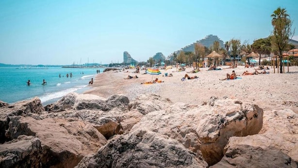 A beach with rocky foreground, people sunbathing, and modern buildings in the background.