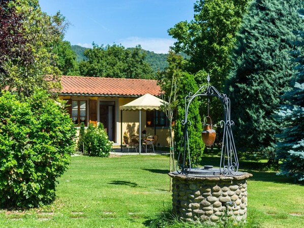 Un puits en pierre avec des éléments métalliques, une maison avec un toit en tuiles et une végétation luxuriante tout autour.