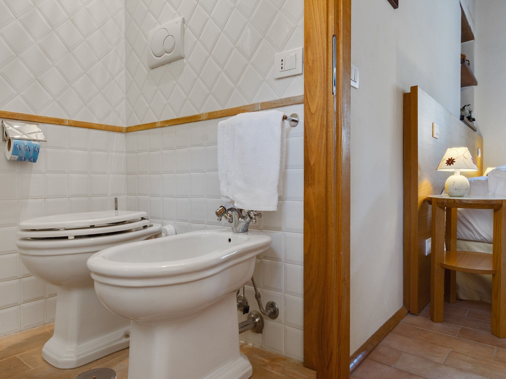 Une salle de bain avec deux toilettes, un lavabo, un porte-serviettes, un distributeur de papier toilette et une chaise en bois.