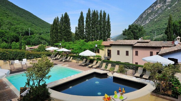 A pool area with lounge chairs and umbrellas, surrounded by greenery and buildings. - Countryside home in Foligno (Foligno)