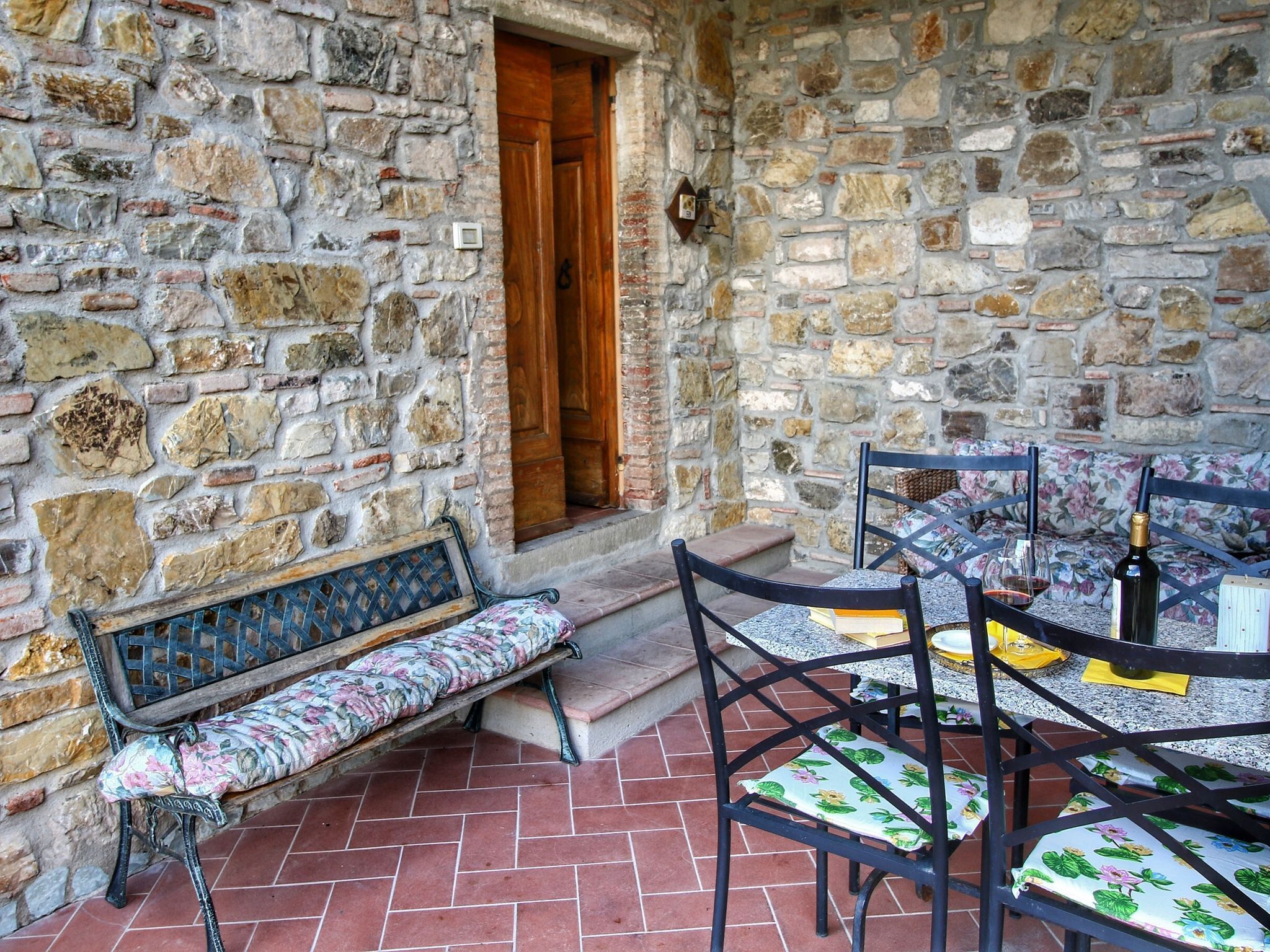 Un espacio al aire libre con paredes de piedra, bancos y sillas, una puerta de madera y suelo de baldosas.