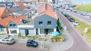Ein modernes Gebäude mit grauer Fassade und rotem Dach, an einer Straßenecke gelegen, mit Parkplätzen und Blick auf das Meer.