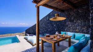 A terrace with a stone wall, a wooden table set with breakfast, and a view of the ocean. - Lithi Luxury Retreat (Santorini)