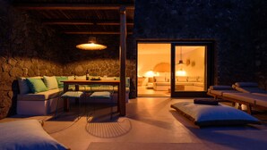 A cozy room with a stone wall, a sofa, a dining table with chairs, and a massage area with chairs. - Lithi Luxury Retreat (Santorini)