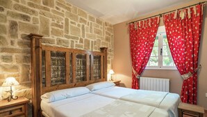 A bedroom with two beds, stone walls, a wooden headboard, red curtains, and a window with a view.