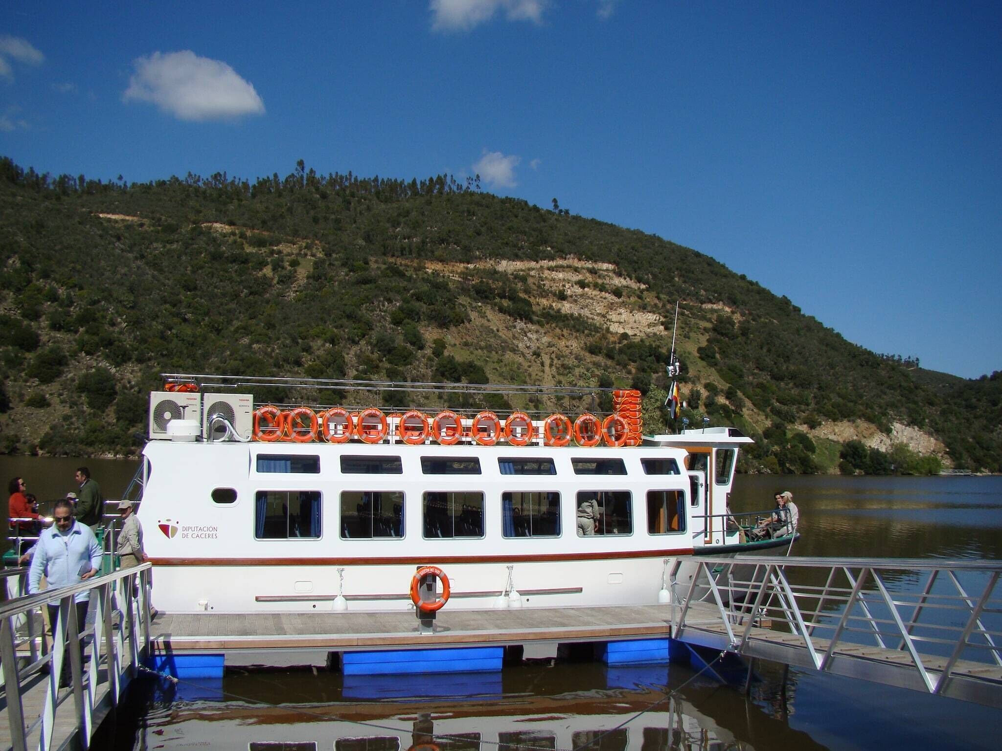 A white boat docked at a pier with lifebuoys on the deck and a mountainous backdrop.