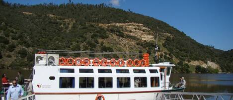 A white boat docked at a pier with lifebuoys on the deck and a mountainous backdrop.