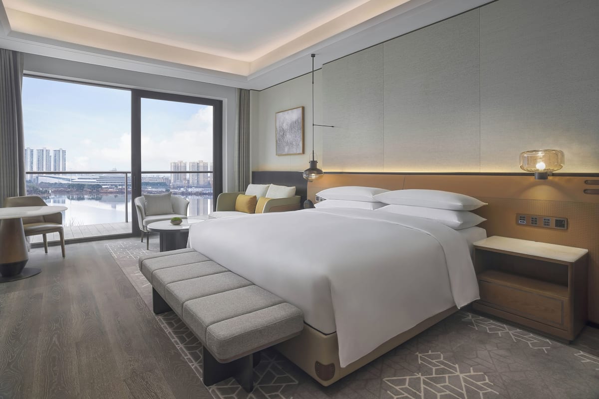 Una moderna camera d'albergo con un letto grande, un comodino, una panca, una scrivania e una sedia. È presente un'ampia finestra con vista su edifici e un ponte.