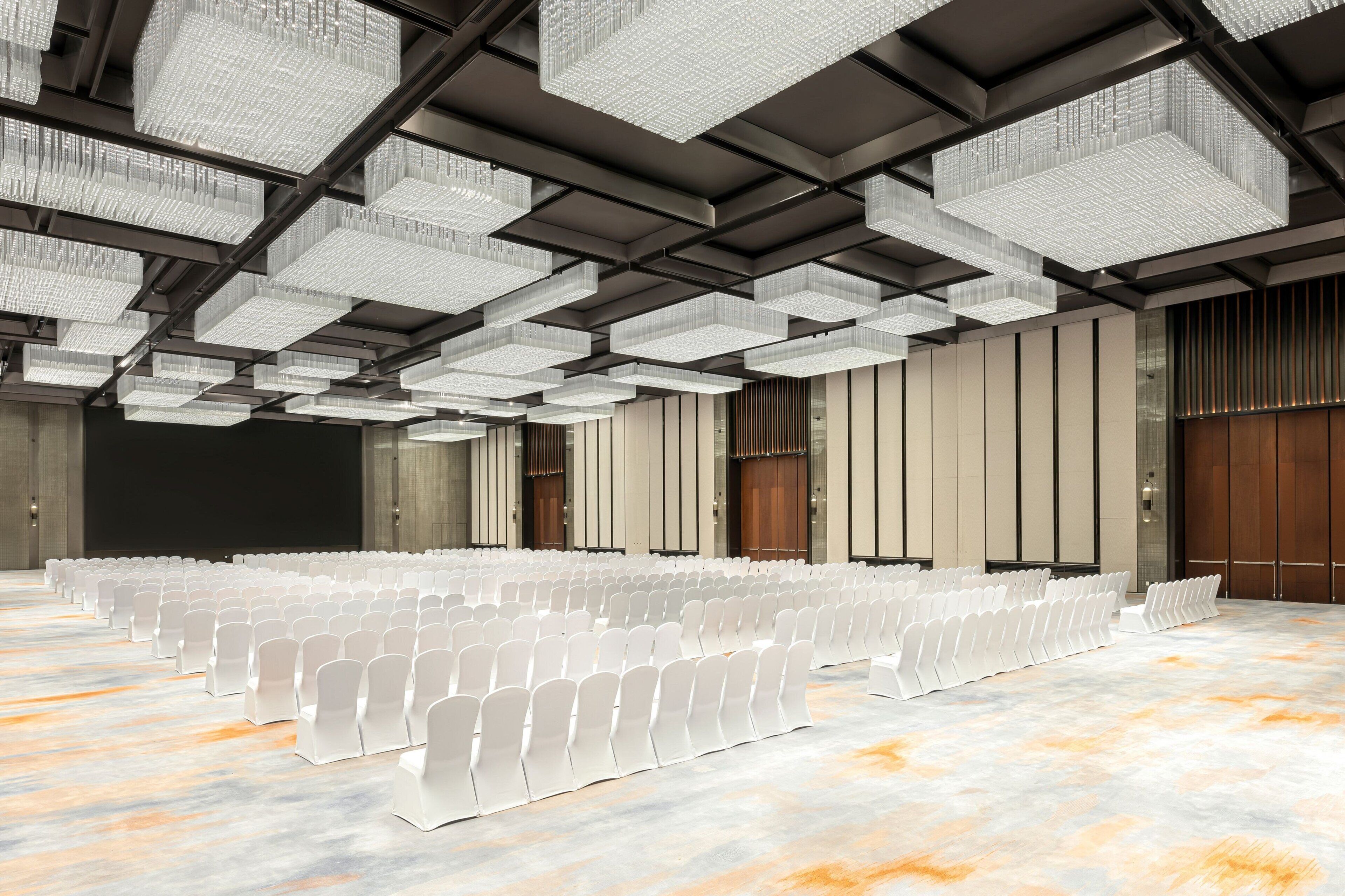 A large hall with rows of white chairs, a high ceiling with hanging lights, and wooden doors.