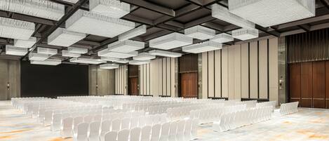 A large hall with rows of white chairs, a high ceiling with hanging lights, and wooden doors.