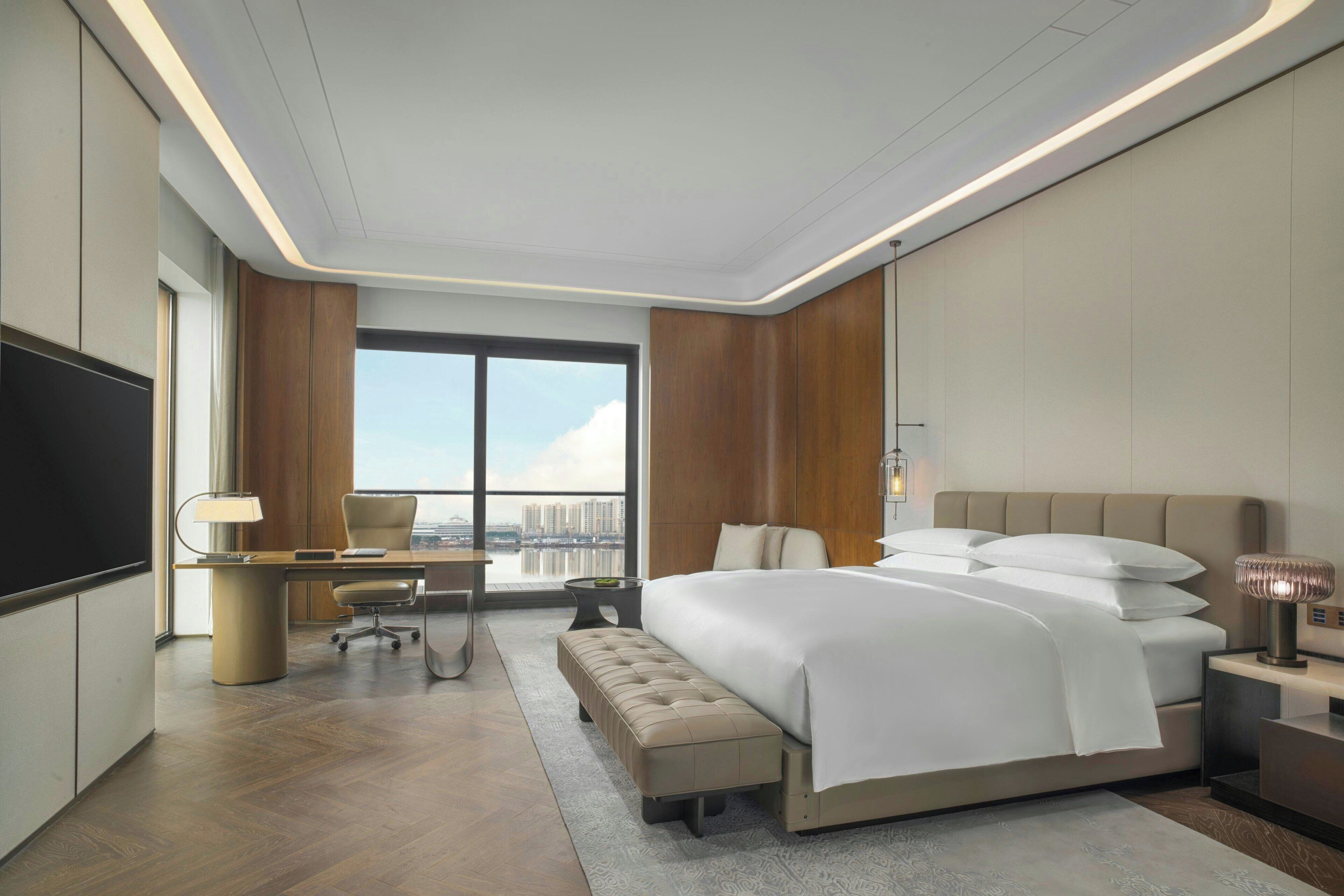 Habitación de hotel moderna con una cama grande, un escritorio, una silla, un televisor y vistas a la ciudad.
