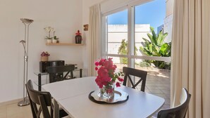 A dining area with a white table, black chairs, and a vase of pink flowers. There is a floor lamp, a shelf with decorative items, and a view of a garden through a window.