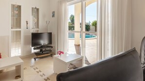 A living room with a TV, a white coffee table, and a sofa. There is a sliding glass door leading to an outdoor area with a pool.