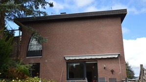 A two-story brick house with a large window and a door, surrounded by a garden with yellow flowers and green shrubs. - Modern Holiday Home in Eifel with Sauna (Stadtkyll)