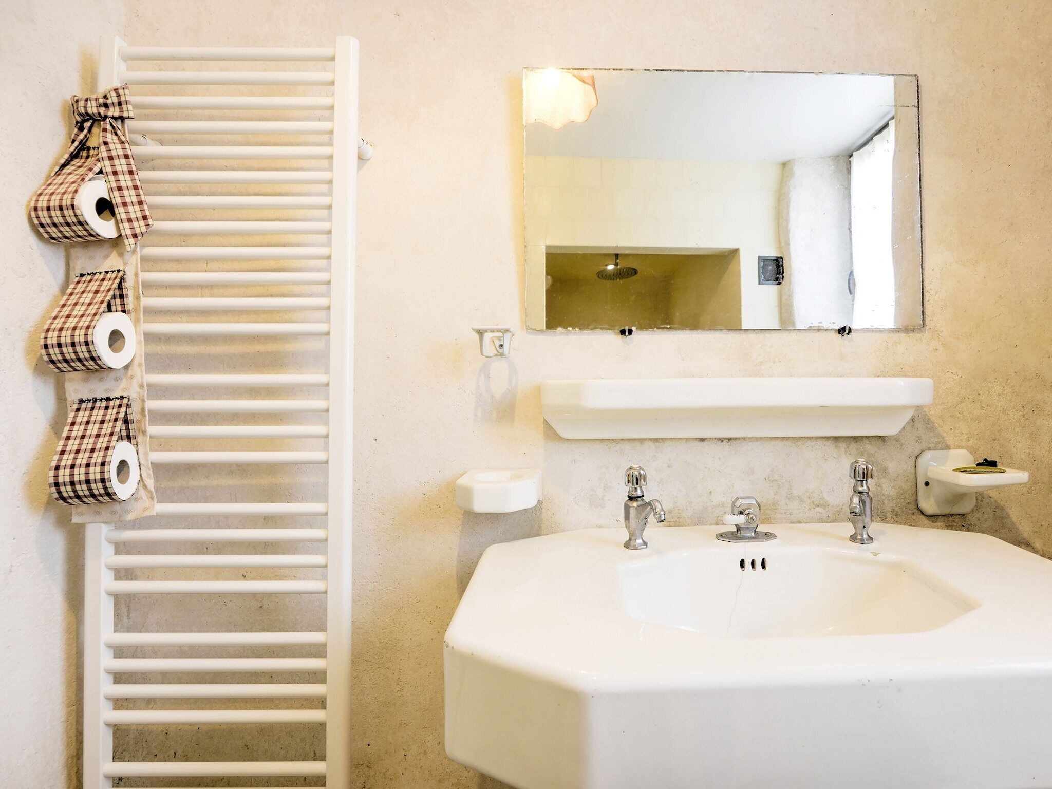 A bathroom with a white sink, two faucets, a mirror above the sink, and a towel rack with rolled towels.