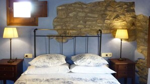 A bedroom with a stone wall, a bed with patterned bedding, two bedside lamps, and a wooden headboard. - Belvilla by OYO Casa Vella (Castigaleu)