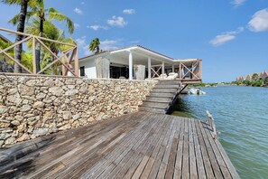 Eine Holzterrasse führt zu einem überdachten Außenbereich mit einer Steinmauer, Palmen und Blick auf das Wasser.
