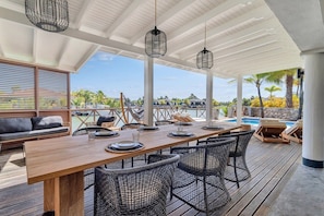 A covered outdoor dining area with a wooden table, black wicker chairs, and hanging pendant lights. - Costa de la Luz with pool on Laguna Marina (Kralendijk)