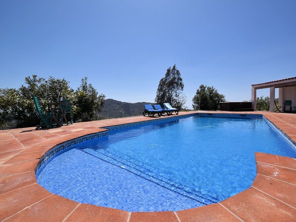Ein Schwimmbecken mit gefliestem Rand, umgeben von einer Terrasse mit Stühlen und einer Pergola.