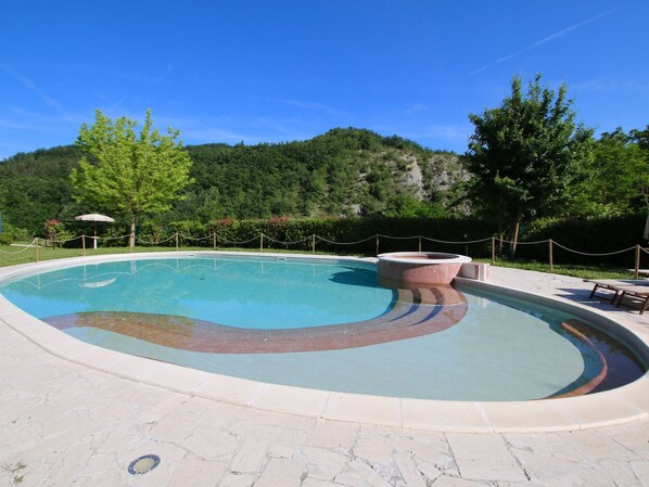 A swimming pool with a hot tub, surrounded by a stone deck, with trees and a hill in the background.