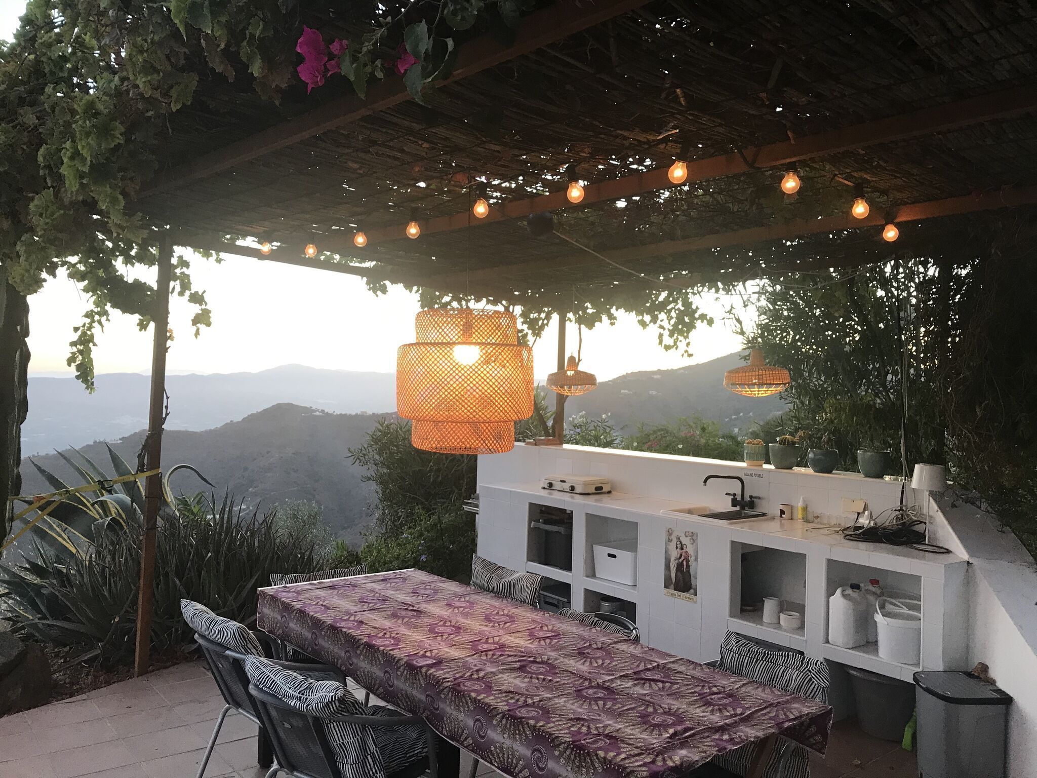 Un área de comedor al aire libre con mesa y sillas, una estación de parilla integrada y lámparas colgantes.