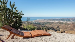 A wooden lounge chair on a gravel surface with a view of a coastal landscape and distant city.