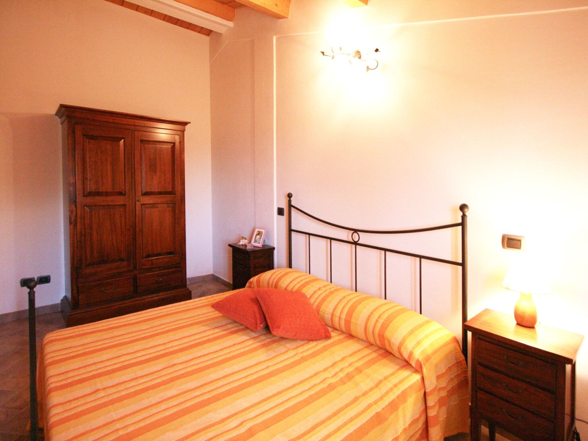A bedroom with a bed, a wooden wardrobe, a bedside table with a lamp, and a wall-mounted light fixture.