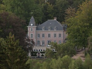 Un grand immeuble à étages, doté d’une façade symétrique, avec une tour centrale surmontée d’une horloge, le tout entouré d’une végétation luxuriante.