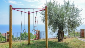 Un'area giochi con altalene e strutture per arrampicata, recintata da una staccionata e con un albero.