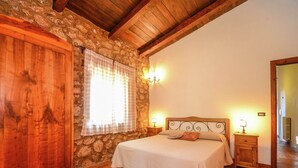 Una camera da letto con parete in pietra, soffitto in legno, un letto con testiera, due comodini con lampade e una finestra con tende.