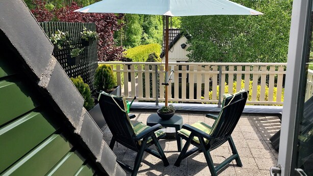 A patio with two chairs and a table under a large umbrella, surrounded by a white fence and greenery.