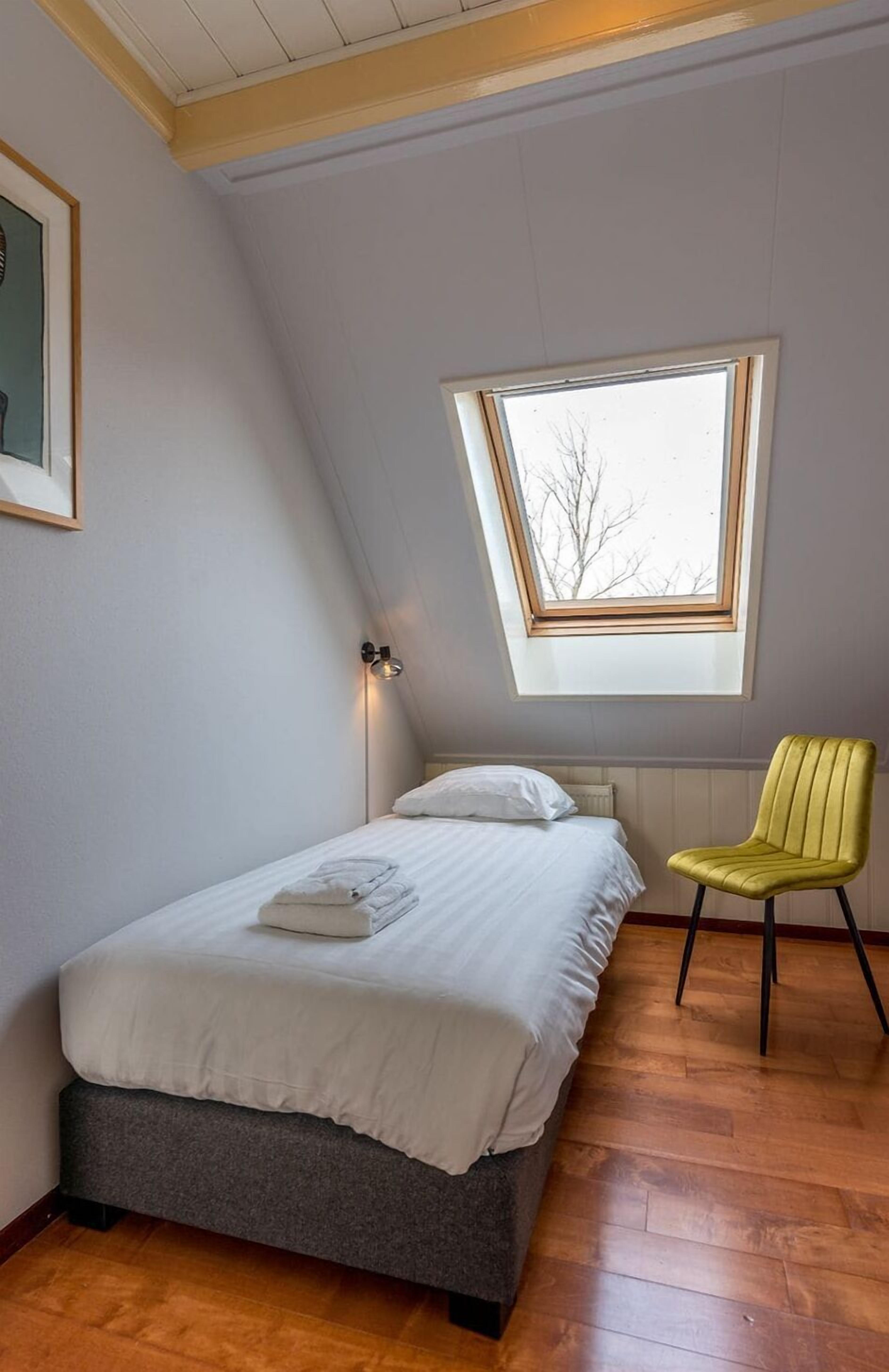 Une chambre à coucher avec un lit, une fenêtre, une chaise et un luminaire fixé au mur.