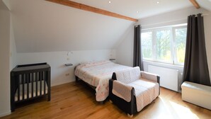 Quarto com teto inclinado, vigas de madeira, cama com colcha, mesa de cabeceira, berço, janela com cortinas e radiador.