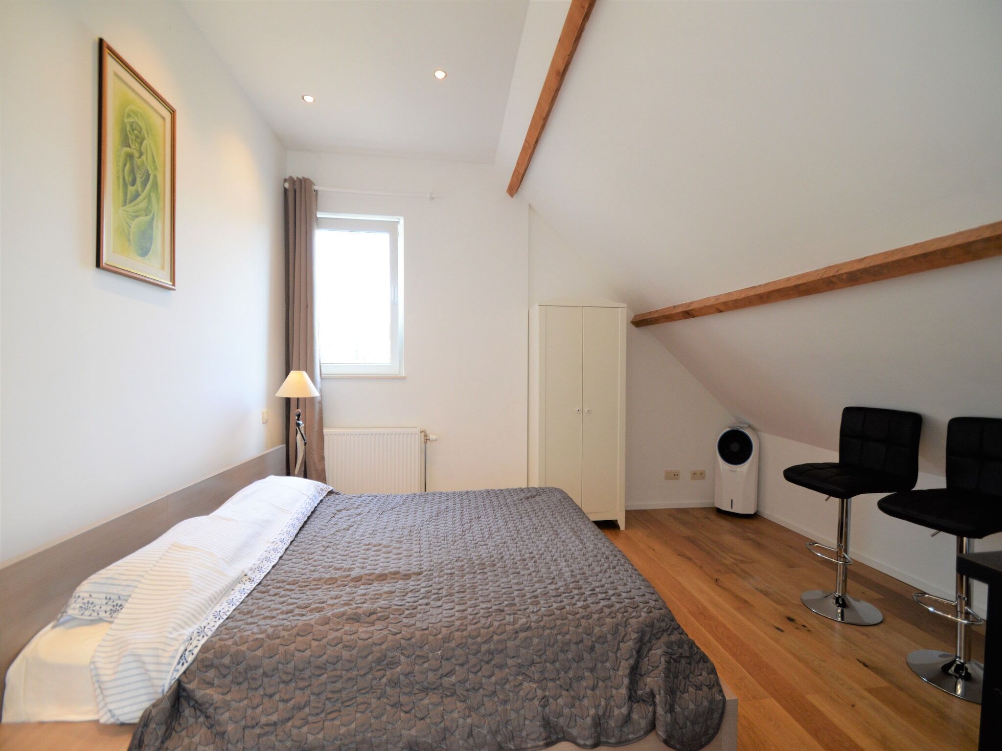 A bedroom with a bed, a window, a radiator, a wardrobe, and two bar stools.