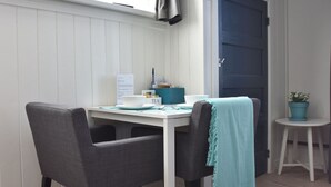 Una habitación pequeña y acogedora con una mesa blanca y dos sillas grises, una puerta azul y una pequeña planta en maceta sobre un taburete blanco.