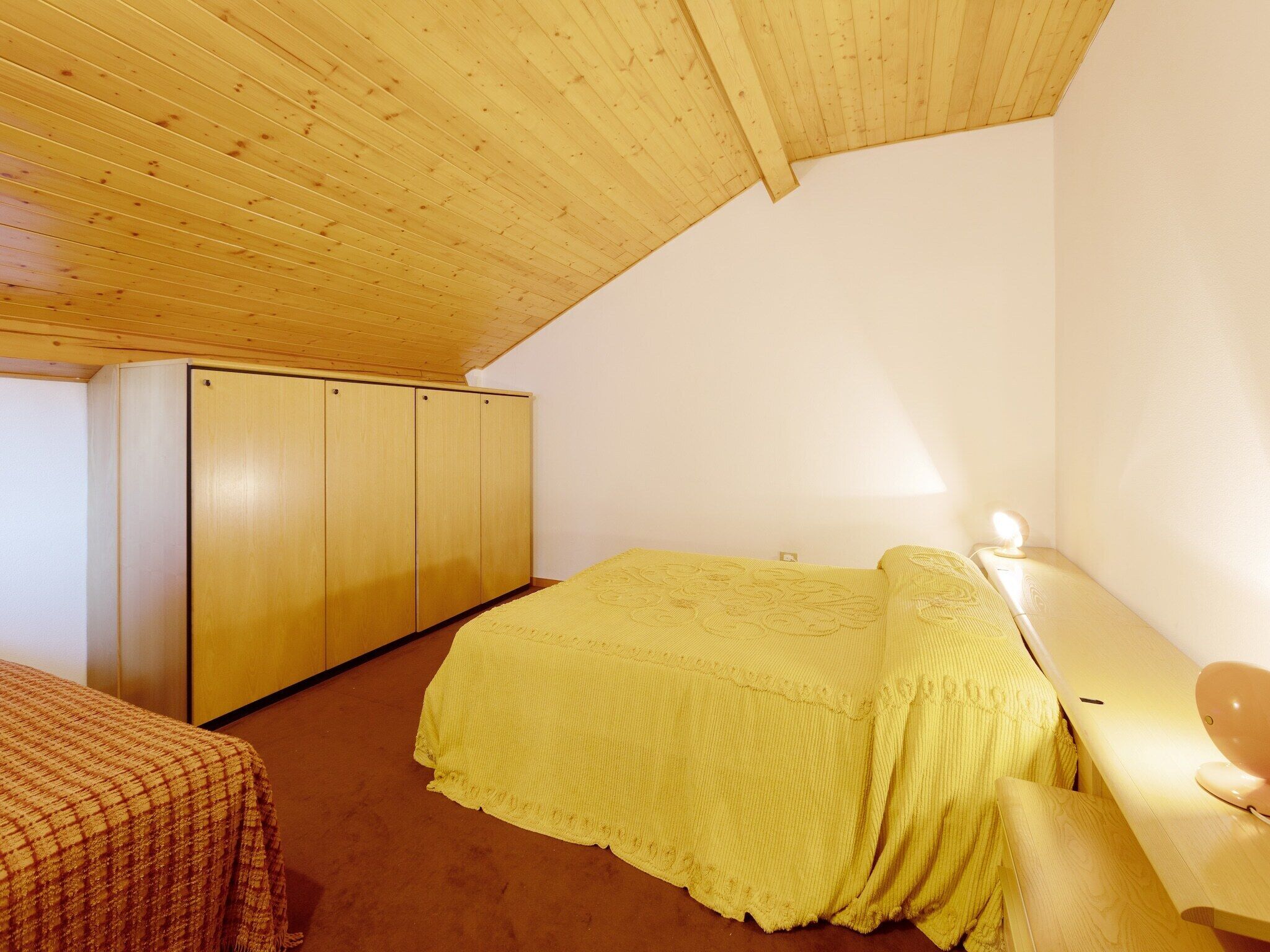 A bedroom with a wooden ceiling, a bed with a yellow bedspread, a wardrobe, and a bedside table with a lamp.