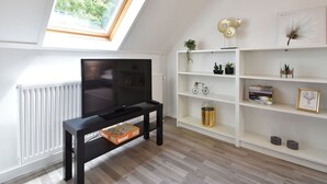 A modern living room with a TV, wooden flooring, white walls, a skylight, and a bookshelf with decorative items. - Modern Holiday Home in Wolphaartsdijk with Private Garden (Wolphaartsdijk)