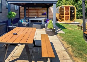 An outdoor patio with a wooden table and benches, a covered seating area, and a sauna.