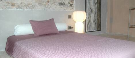 A bed with a pink quilted bedspread, a white pillow, and a purple pillow. There is a wall with a stone texture and a painting above the bed.