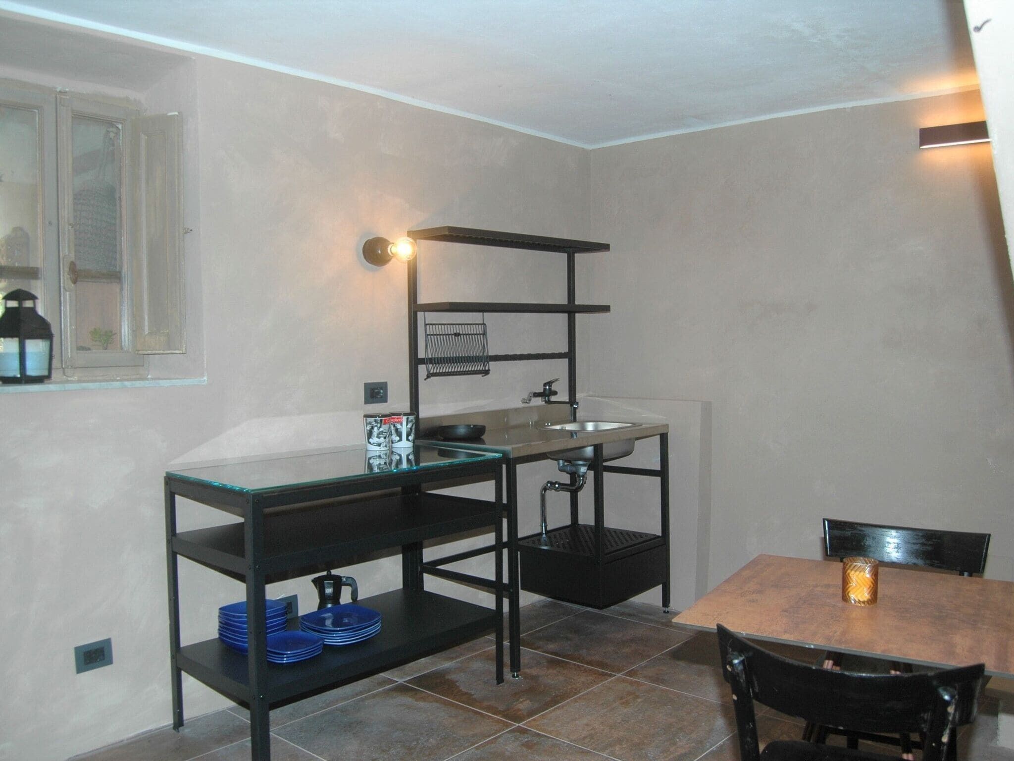Une cuisine compacte avec une table à plateau en verre, des étagères en métal noir, un évier et une fenêtre ornée d’une lanterne.