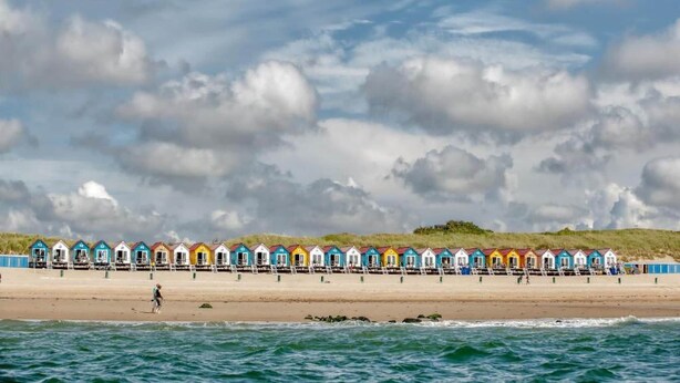 Una hilera de cabañas de playa coloridas a lo largo de una playa arenosa, con vista al mar y un cielo nublado.