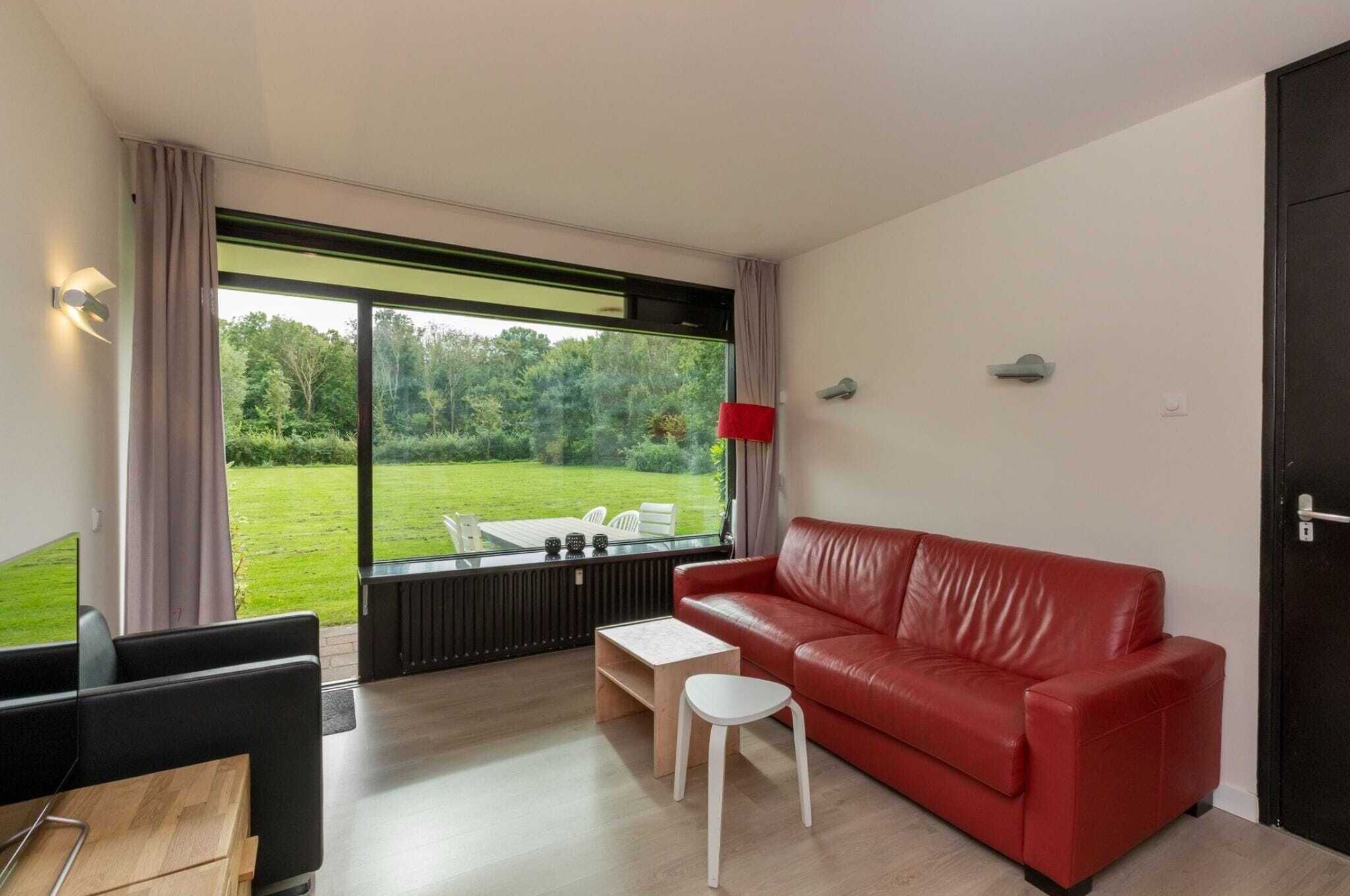Un salon moderne avec un canapé en cuir rouge, une petite table blanche et une table basse en verre. Une grande fenêtre offre une vue sur une pelouse verdoyante et des arbres.