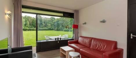 Un salon moderne avec un canapé en cuir rouge, une petite table blanche et une table basse en verre. Une grande fenêtre offre une vue sur une pelouse verdoyante et des arbres.