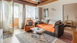 Ein gemütliches Wohnzimmer mit einer Couch, einem kleinen Tisch und einem Sessel. Ein Fenster mit Vorhängen, ein Deckenventilator und eine Wandleuchte sind ebenfalls vorhanden.