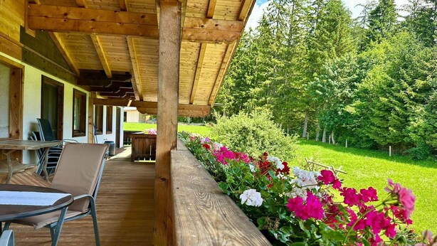 Een overdekt buitengebied met een houten terras, een tafel met stoelen en een bloembed met roze en witte bloemen.