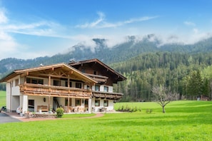 Un edificio de estilo chalete con balcón de madera, rodeado de un paisaje verde exuberante y montañas al fondo.