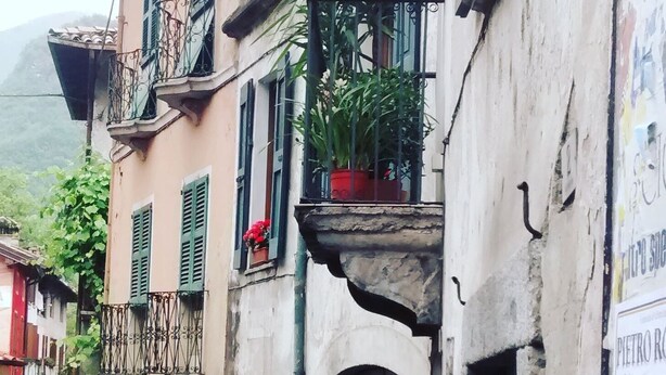 A narrow street with old buildings, balconies with plants, and a door with a window above it. - Ostello Sociale Borgo Venno (Lavenone)
