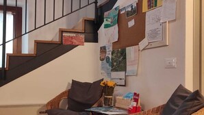 A staircase with a black railing, a bulletin board with various papers and notices, wicker chairs, a small table with a vase of flowers, and a red box on the table. - Ostello Sociale Borgo Venno (Lavenone)